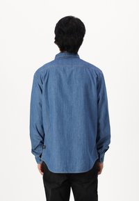 Camicia di jeans blu medio, maniche lunghe, dettaglio del giogo posteriore, orlo curvo, con polsini a bottoni e spacchi laterali, realizzata in tessuto di cotone leggero.