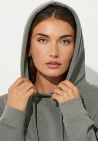 Grijze hoodie met een trekkoordkap, ribgebreide manchetten en zachte stof. Het model houdt de kap vast, waardoor een strakke, minimalistische vormgeving zichtbaar wordt.