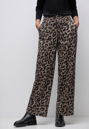 Kvinde iført vidde bukser med brun og sort leopardprint, sort top, sorte ankelstøvler, hænderne i lommerne, stående foran en ensfarvet baggrund.