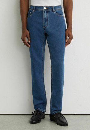 Mand iført blå straight-leg jeans og sorte snørebåndssko, stående mod en lyset struktureret væg.