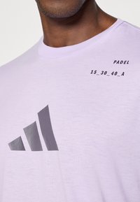 Liliowa koszulka z krótkim rękawem wykonana z miękkiego materiału, z ciemnoszarym nadrukiem logo i napisem "PADDEL 15_30_40_A" z przodu.