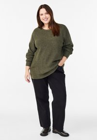 Oliivivihreä neulepaita rentolla leikkauksella ja varmistetuilla harteilla, yhdistettynä löysiin mustiin housuihin ja nilkkureihin. Kaksi etutaskua.