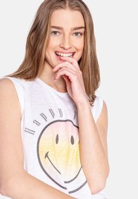 Wit mouwloos T-shirt met een grafische afbeelding van een lachend gezicht; zachte textuur; bevat de tekst "Happy Vibes" in een speelse lettertype boven de grafiek.