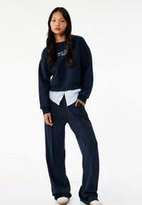 Sudadera navy recortada con texto bordado, superpuesta sobre una camisa de rayas azul claro, combinada con pantalones de pierna ancha navy y zapatillas blancas.