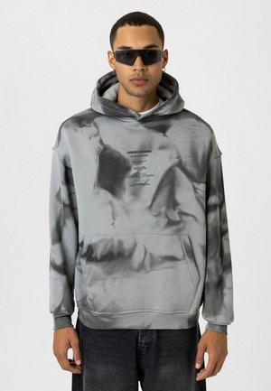 Grijze tie-dye hoodie met een voorzak, voorzien van een bedrukt logo en tekst. De stof lijkt soepel met een comfortabele fit en een grote capuchon.