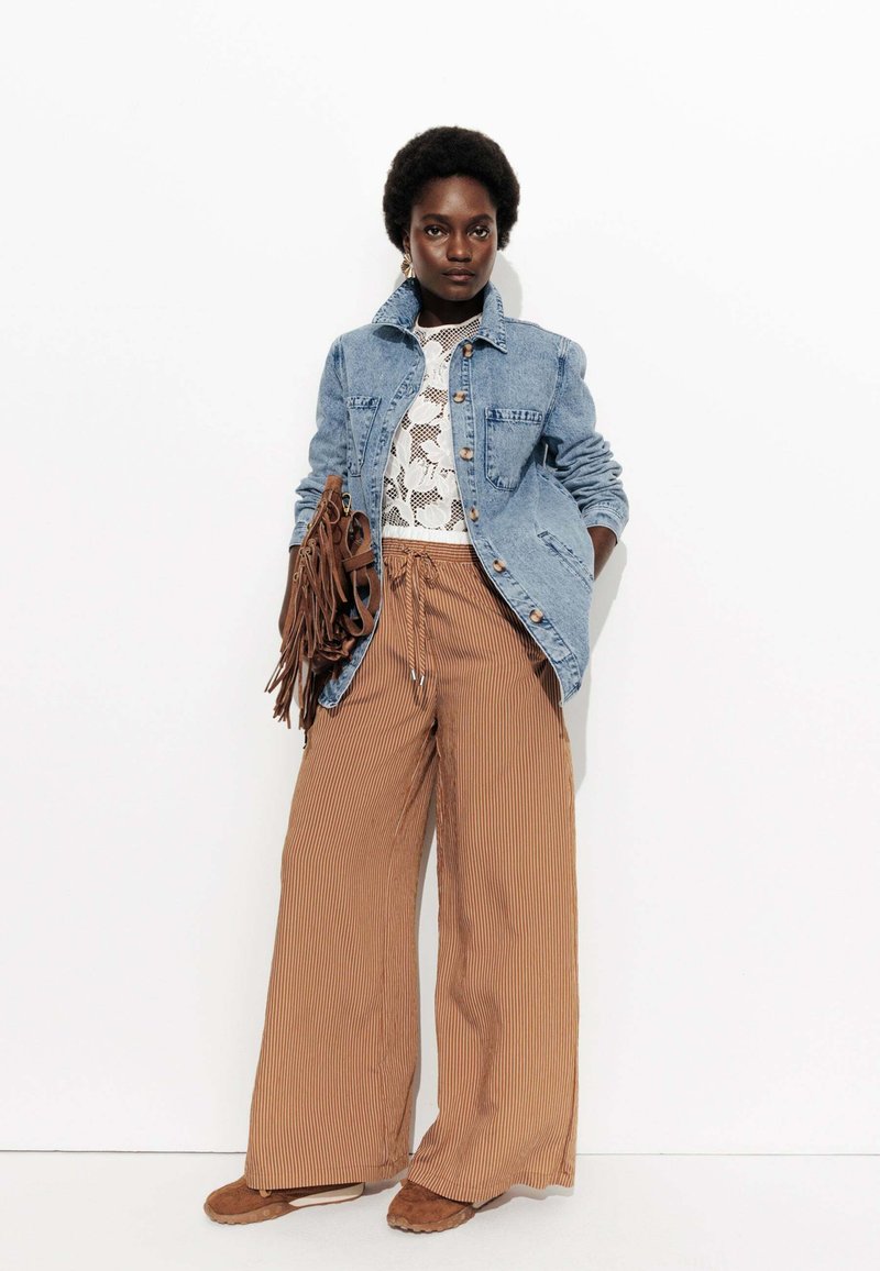 Veste en denim avec fermeture à boutons, associée à un top en dentelle blanc et à un pantalon large à rayures brunes. Comprend un sac à franges marron et des chaussures beiges.