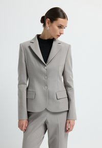 Blazer gris clair avec une silhouette ajustée, deux poches avant, trois boutons argentés et des revers crantés. Porté par-dessus un top noir texturé.