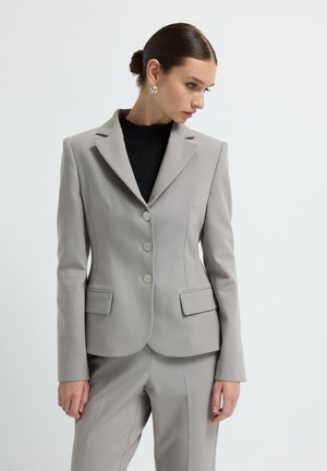 Light grey blazer with a fitted silhouette, two front pockets, three silver buttons, and notched lapels. Worn over a black textured top.