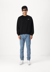Svart hettegenser med rund hals og liten logo, kombinert med lyseblå jeans og hvite joggesko. Modell med mørke solbriller.