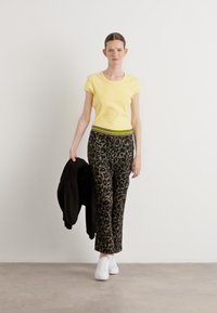 Geel T-shirt met korte mouwen, zwarte broek met luipaardprint en groene streep in de taille, witte sneakers en een zwart jasje dat over de linkerarm hangt.