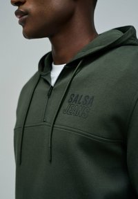 Sudadera con cremallera de color verde oscuro, hecha de un tejido suave, con un logo bordado "SALSA JEANS" en el pecho y un diseño de bolsillo tipo canguro.