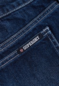 Tissu en denim bleu foncé présentant des coutures visibles et une étiquette noire avec "SUPERDRY" en blanc, accompagnée d'un logo en forme de blason.