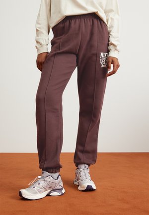 Personne portant un pantalon de jogging Nike marron foncé et des chaussures de sport blanches, debout sur un sol marron sur un fond uni.