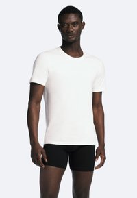 Hvid bomulds T-shirt med rund hals, korte ærmer og en figursyet design. Modellen har sorte shorts på. Glat tekstur, minimal detaljering.