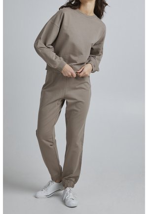 Person wearing taupe-colored sweatshirt and matching jogger pants with white sneakers, adjusting the sweatshirt hem against a plain background.