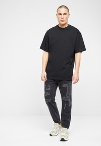 Camiseta negra de gran tamaño combinada con jeans negros desgastados que presentan parches deshilachados. El modelo lleva zapatillas de charol beige claro. Fondo simple.