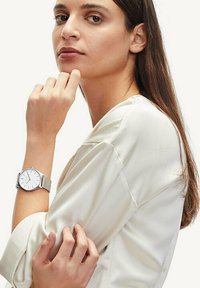 Silberne Armbanduhr mit einem Netzarmband, rundem weißen Zifferblatt und silbernen Stundenmarkierungen. Das Modell trägt eine hellcremefarbene Bluse mit lockerer Passform.