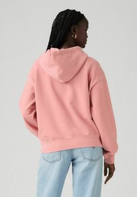 Sudadera rosa con capucha, de corte relajado y puños acanalados. Combinada con jeans de color azul claro, mostrando un estilo casual y cómodo.