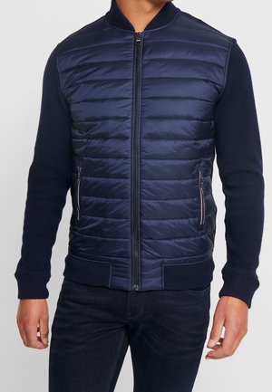 Mann trägt eine marineblaue gesteppte Jacke mit Reißverschluss, gestrickten Ärmeln und Kragen, kombiniert mit dunklen Jeans, steht vor einem einfarbigen Hintergrund.