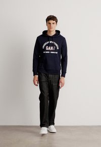 Granatowa bluza z kapturem z białym napisem logo, w połączeniu z ciemnoniebieskimi dżinsami i białymi sneakersami. Model stoi na jednolitym tle.