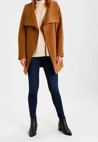 Brauner Wollmischungsmantel mit weitem Kragen, getragen über einem hellen Pullover, kombiniert mit dunklen Jeans und Stiefeletten. Glatte Textur und lässige Passform.