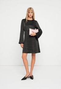 Svart satindress med lange bel sleeves, en tettsittende midje og en utsvingt skjørt. Modellen holder en myk rosa clutch og har på seg svarte spisse hæler.