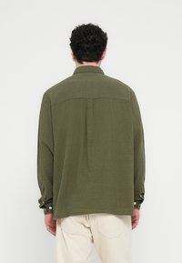 Vīrietis ar īsiem cirtainiem matiem, ģērbies olīvu zaļā garroku kreklā un gaiši bēšās biksēs, nofotografēts no aizmugures pret vienkāršu fonu.