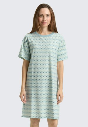 Vrouw staat, draagt een loszittende, jurk met korte mouwen tot op de knieën, met een lichtblauw geometrisch patroon op een bleke achtergrond.