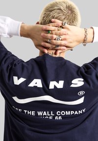 Camiseta de la marina con un gran logo en blanco de "VANS" y texto. El modelo tiene el cabello corto y rubio, y múltiples anillos en las manos, con los brazos cruzados detrás de la cabeza.