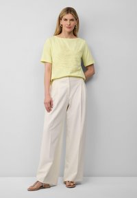 Kobieta w jasnożółtej bluzce z krótkim rękawem z haftem liści, w kremowych szerokich spodniach, w złotych kolczykach w kształcie kół i płaskich sandałach, stojąca przed szarym murem.