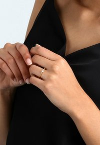 Goldring mit einem filigranen Design, der drei klare Steine zeigt. Die Hände liegen natürlich vor einem schwarzen Stoffhintergrund.