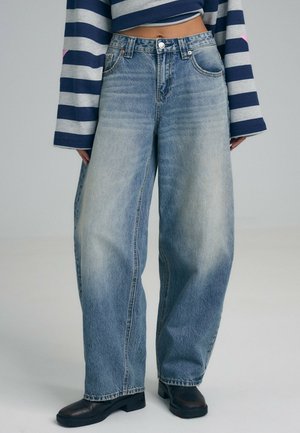 Le mannequin porte un jean ample bleu clair délavé, un pull bleu marine à rayures et des chaussures noires, sur un fond de studio uni.