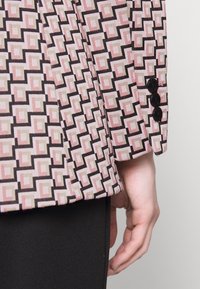 Sako s geometrickým vzorem v růžové, černé a béžové barvě, vyrobené z texturované látky, s černými knoflíky na rukávech.