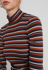 Top de cuello alto de mangas largas y con canalé, que presenta rayas horizontales en azul marino, óxido, crema y rojo. Diseño ajustado con una textura flexible.