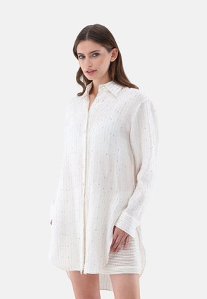Camicia bianca con righe verticali e sottili punti metallici. Presenta una chiusura con bottoni, maniche lunghe e una vestibilità leggermente ampia.