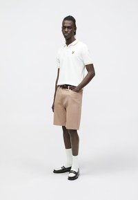 Vit pikétröja med gula detaljer, beige randiga shorts, brunt bälte, vita strumpor och svarta skor. Modellen står mot en enfärgad bakgrund.