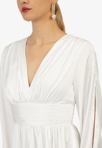 Witte jurk met een diepe V-hals,Gathered taille en lange mouwen met openingen. Gladde satijnen stof met een zachte glans.