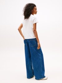 Weißes T-Shirt und lockere, weit geschnittene blaue Jeans. Die Jeans haben Gesäßtaschen und sind aus Denim hergestellt. Das Modell hat lockige Haare und wird von der Seite gesehen.
