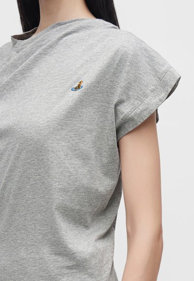 Γυναίκα που φοράει γκρι κοντομάνικη μπλούζα με μικρό πολύχρωμο κεντημένο λογότυπο στο αριστερό μέρος του στήθους.