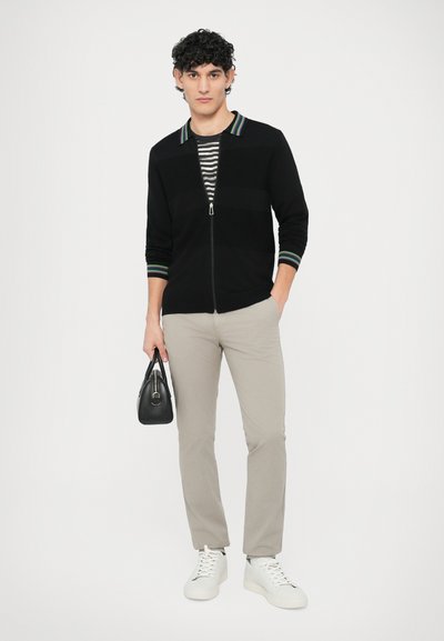 Čierny pletený sveter so zipsom a pruhovaným golierom, sivé nohavice a biele tenisky. Modelka drží malú čiernu tašku a stojí pred jednoliatym pozadím.