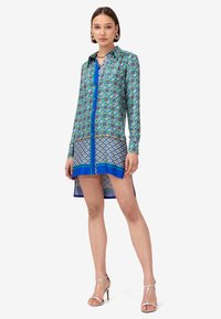 Abito camicia a maniche lunghe con motivi verdi, blu e bianchi; presenta una chiusura con bottoni, accenti blu a contrasto e un orlo curvo.