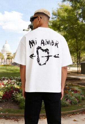 Mann mit kurzen blonden Haaren und Sonnenbrille trägt ein weißes T-Shirt mit schwarzem "Mi Amor"-Text und Herz mit Pfeil Grafik, steht im Park.