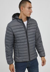 Siva puffer jakna s kapuco, z vodoravnimi kvadratnimi vzorci, sprednjim zadrgo in črnimi akcenti. Nosi se preko belega majice.