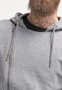 Sudadera con capucha gris con cremallera, de textura suave, que incluye capucha con cordón y detalle de cremallera metálica. Se ve una camiseta negra debajo.