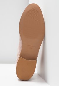 Semelle d'une chaussure en daim beige inclinée contre un mur d'angle blanc, montrant une semelle en caoutchouc texturée et la taille 37 marquée "Fabriqué en Espagne".