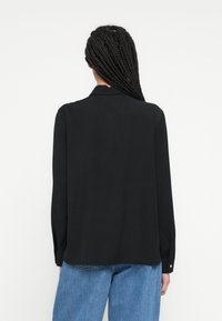 Fekete hosszú ujjú ing sima anyagból, gallérral és gombos mandzsettákkal, laza szabással és finom hátulsó díszítéssel.