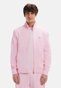 Veste zippée rose clair en molleton doux, avec un col montant, des poches avant et un petit logo sur la poitrine.