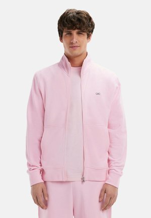 Veste zippée rose clair en molleton doux, avec un col montant, des poches avant et un petit logo sur la poitrine.