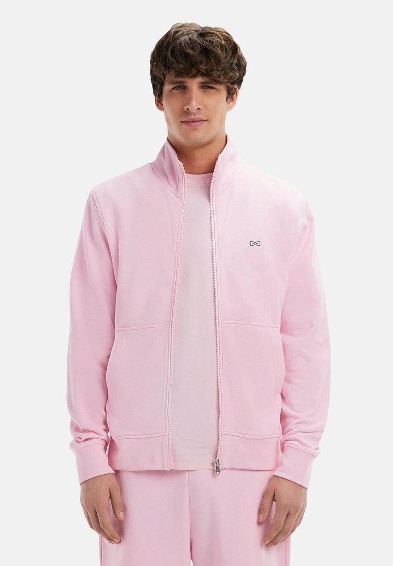 Veste zippée rose clair en molleton doux, avec un col montant, des poches avant et un petit logo sur la poitrine.