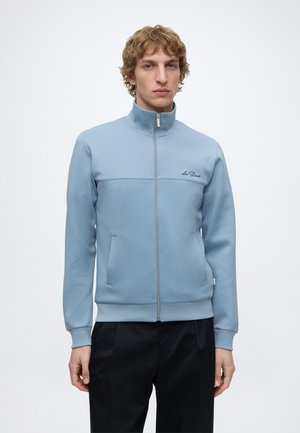 Joven con cabello rizado que lleva una chaqueta azul claro con cremallera y bolsillos y pantalones negros, de pie frente a un fondo blanco liso.
